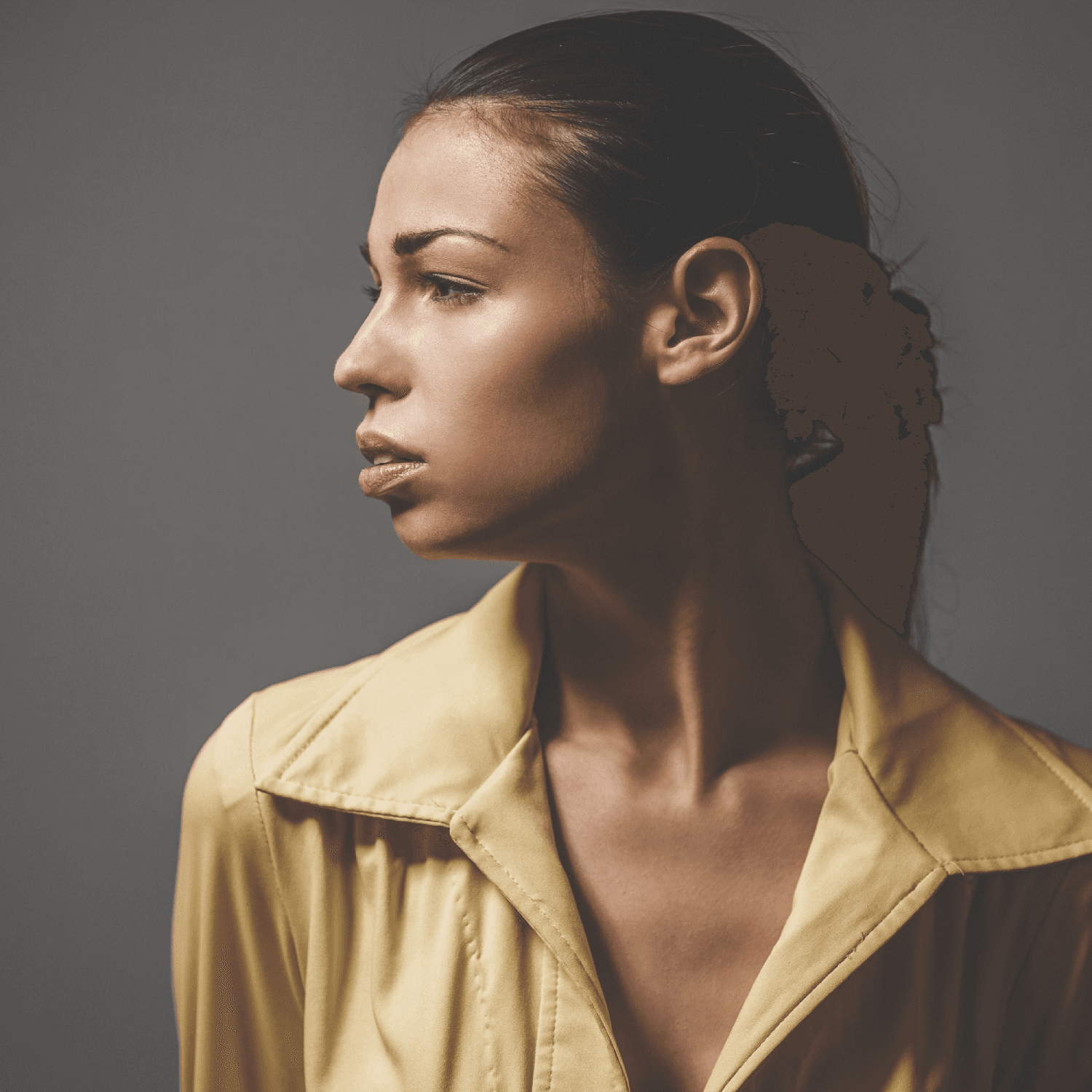 Woman in a pale yellow shirt, dark hair pulled tightly back into a low bun, looking to the side against a grey backdrop. Woman in a pale yellow shirt, dark hair pulled tightly back into a low bun, looking to the side against a grey backdrop.