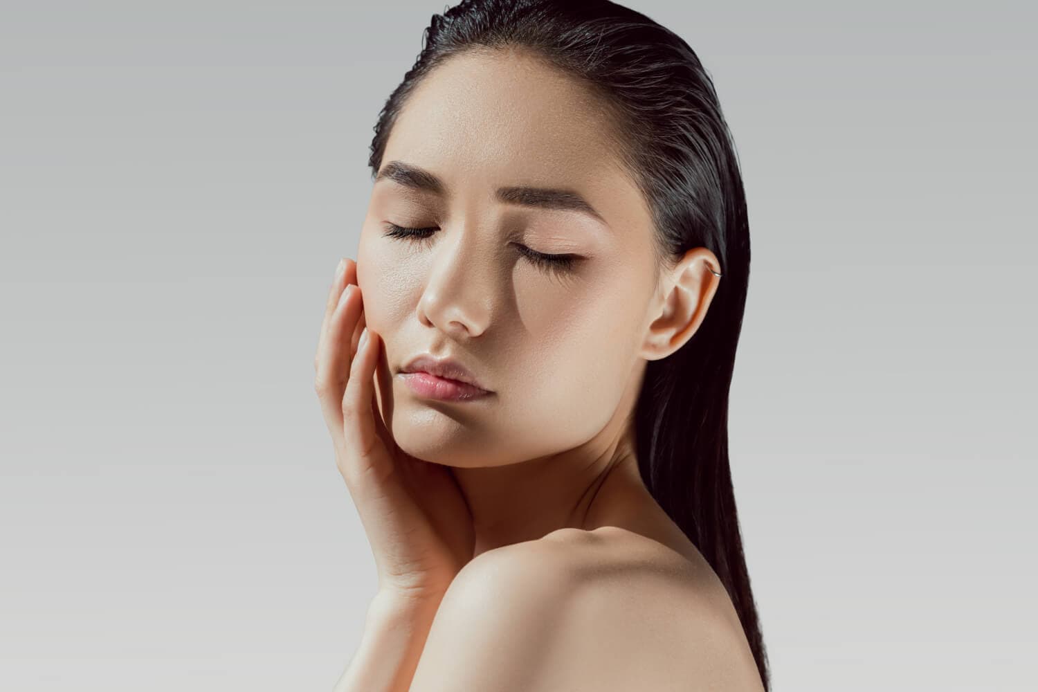Woman with slicked-back dark hair and bare shoulders, eyes closed, resting a hand against her cheek on a grey studio background. Woman with slicked-back dark hair and bare shoulders, eyes closed, resting a hand against her cheek on a grey studio background.