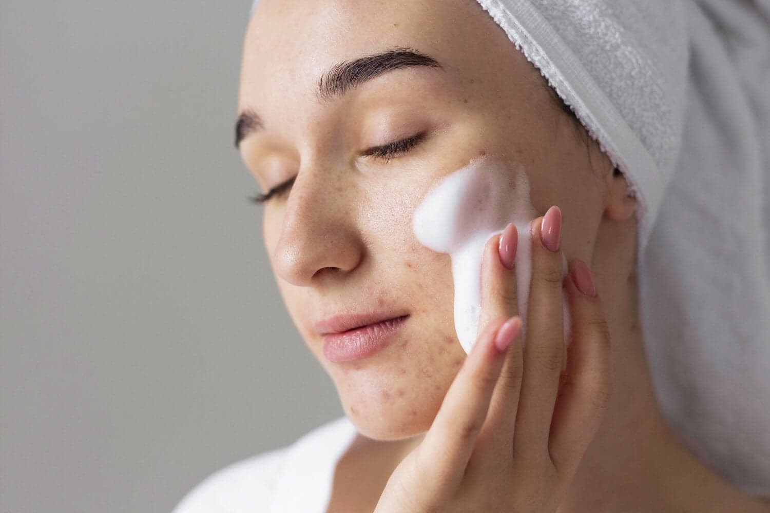 Woman with a white towel turban applying foaming cleanser to her cheek, blemishes and freckles visible on her skin. Woman with a white towel turban applying foaming cleanser to her cheek, blemishes and freckles visible on her skin.