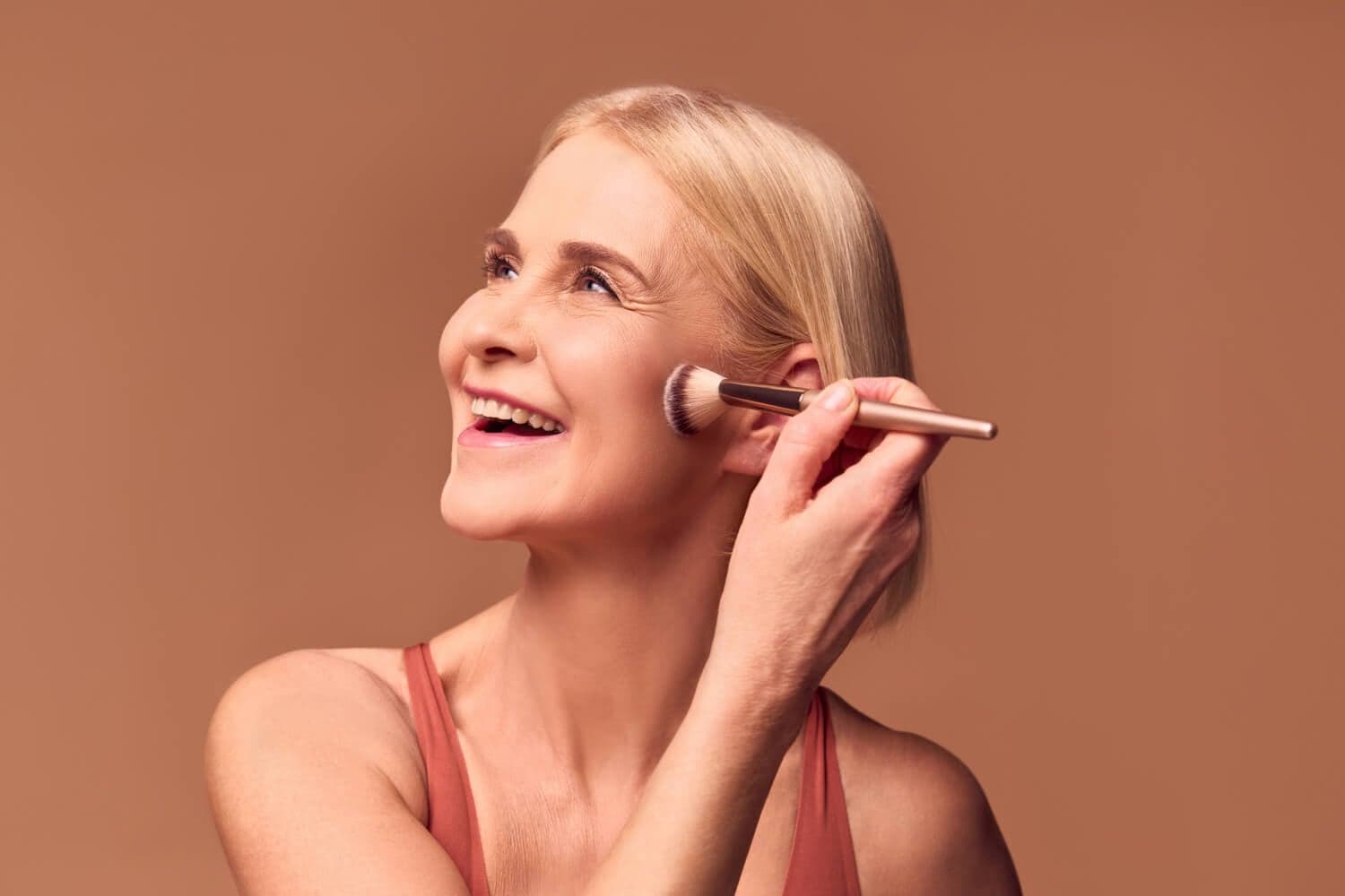 Smiling blonde woman in a rust tank top sweeping a gold-handled powder brush along her cheekbone against a warm brown backdrop. Smiling blonde woman in a rust tank top sweeping a gold-handled powder brush along her cheekbone against a warm brown backdrop.