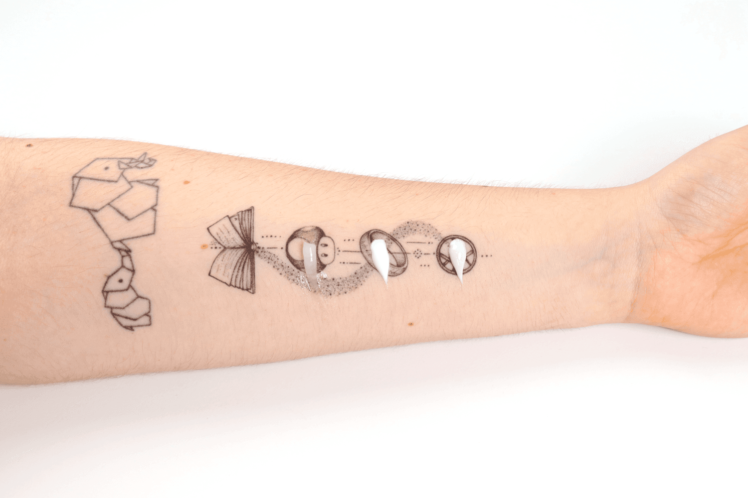 Fine-line black tattoos of origami birds and an open book on a pale forearm, with three swatches of clear gel and white cream smeared over the ink. Fine-line black tattoos of origami birds and an open book on a pale forearm, with three swatches of clear gel and white cream smeared over the ink.