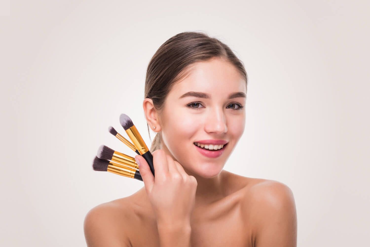 Smiling woman holding a fanned-out set of gold-ferruled makeup brushes against her cheek on a pale grey background Smiling woman holding a fanned-out set of gold-ferruled makeup brushes against her cheek on a pale grey background