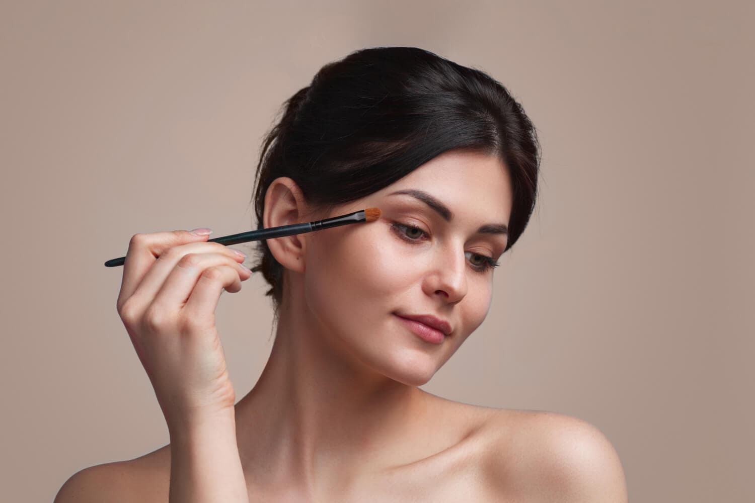 Dark-haired woman with bare shoulders applying soft brown eyeshadow to her eyelid with a flat brush against a beige backdrop. Dark-haired woman with bare shoulders applying soft brown eyeshadow to her eyelid with a flat brush against a beige backdrop.