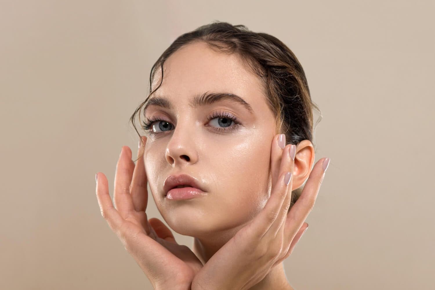 Woman with slicked-back hair framing her dewy, glossy bare face with both hands against a beige backdrop. Woman with slicked-back hair framing her dewy, glossy bare face with both hands against a beige backdrop.