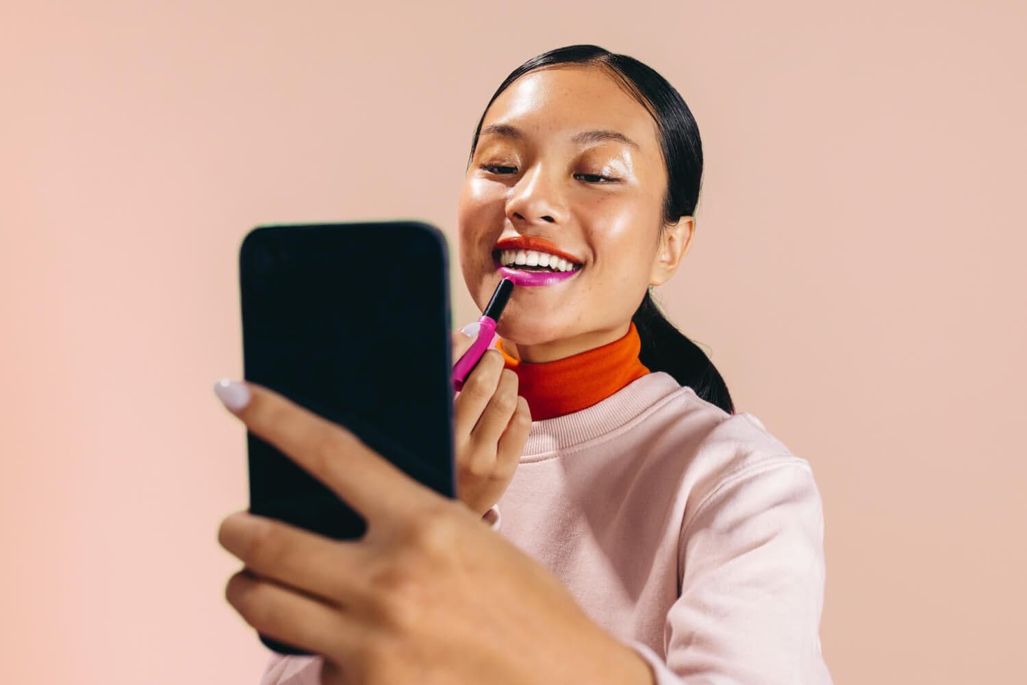 Smiling woman applying bright pink lipstick while looking at her phone screen, against a soft pink backdrop. Smiling woman applying bright pink lipstick while looking at her phone screen, against a soft pink backdrop.