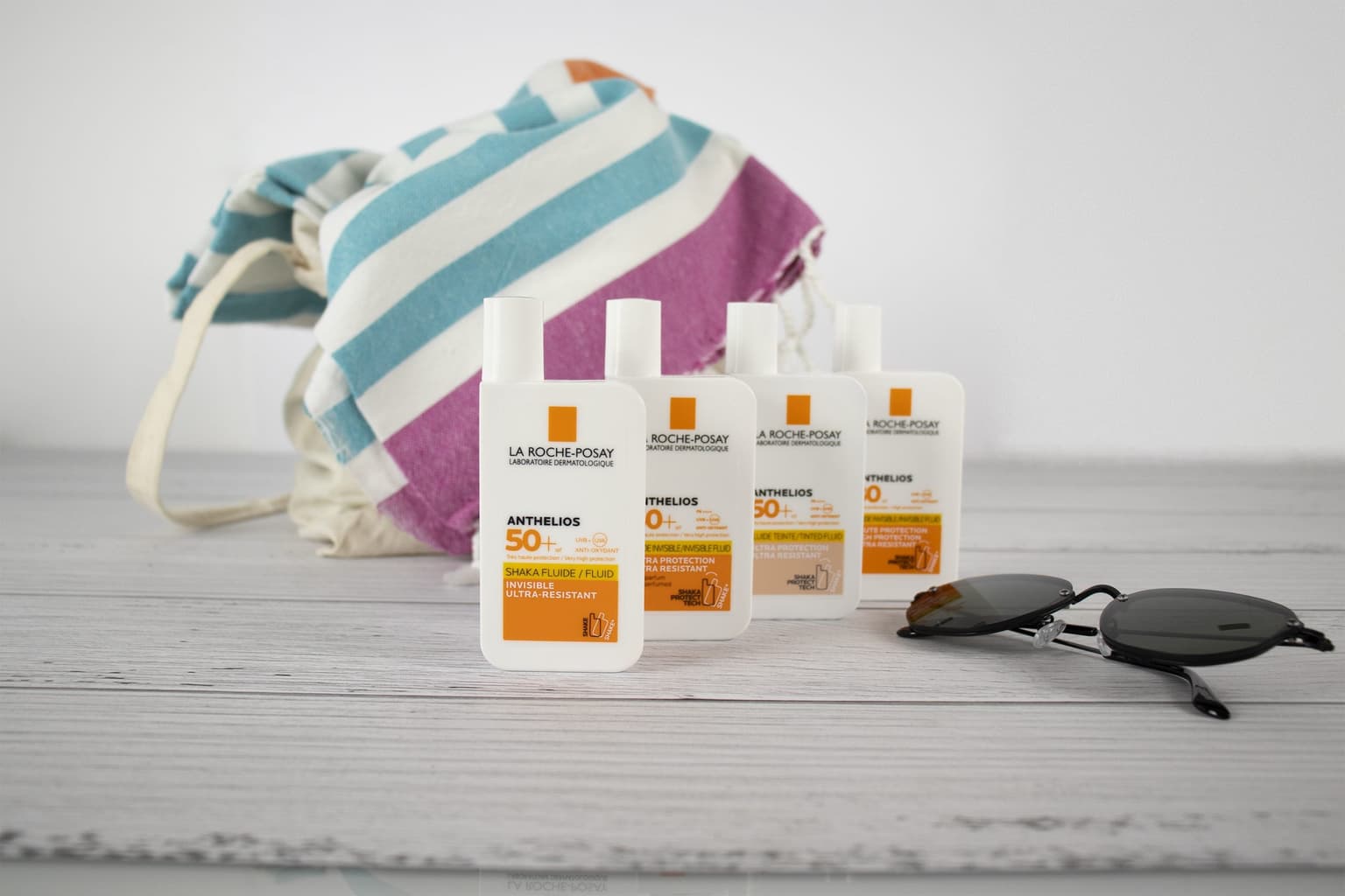 Four La Roche-Posay Anthelios SPF 50+ Shaka Fluid bottles, invisible and tinted, beside sunglasses and a striped beach bag. Four La Roche-Posay Anthelios SPF 50+ Shaka Fluid bottles, invisible and tinted, beside sunglasses and a striped beach bag.