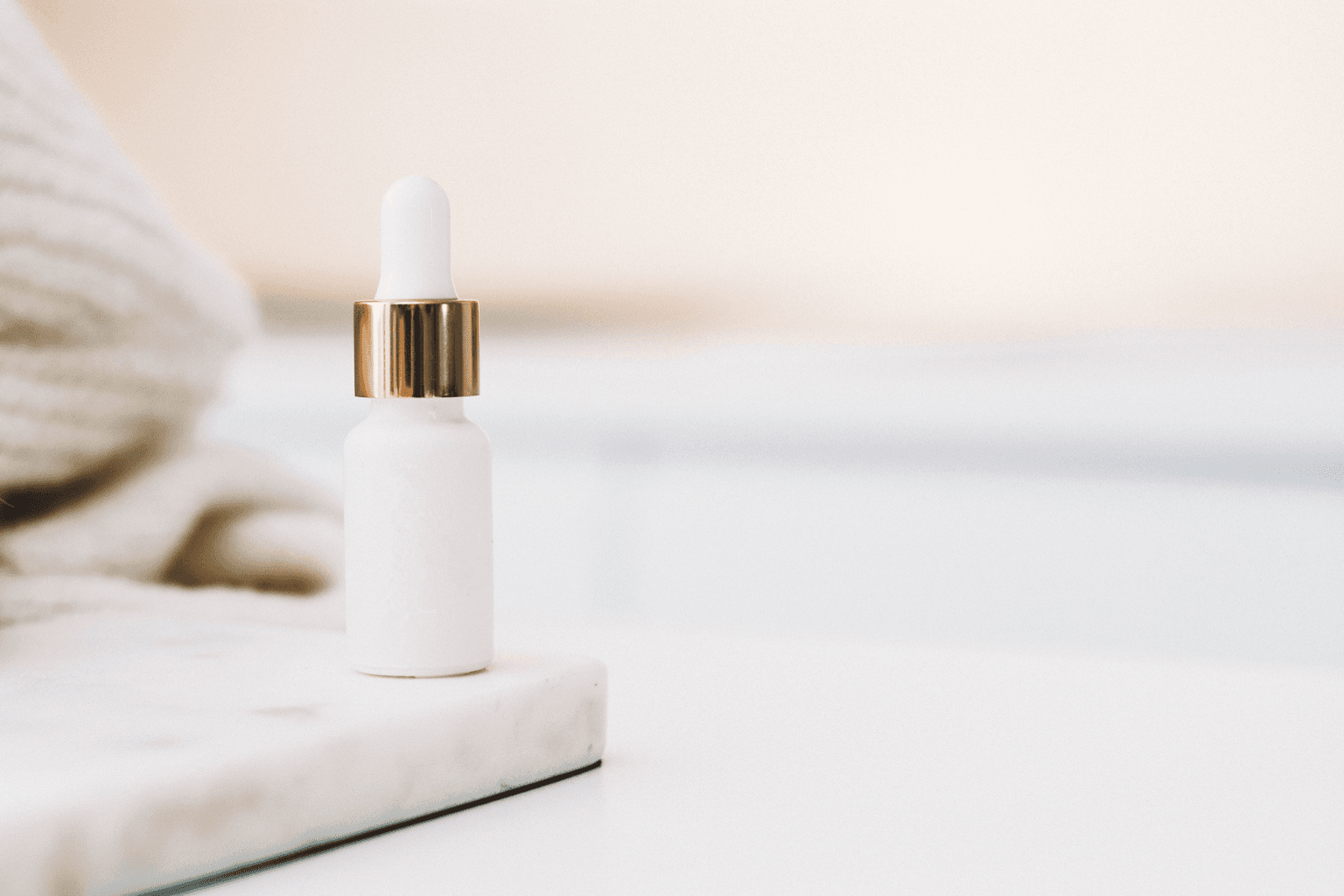 Unlabelled white serum bottle with a gold collar and white dropper cap, standing on a marble slab beside a knitted throw. Unlabelled white serum bottle with a gold collar and white dropper cap, standing on a marble slab beside a knitted throw.