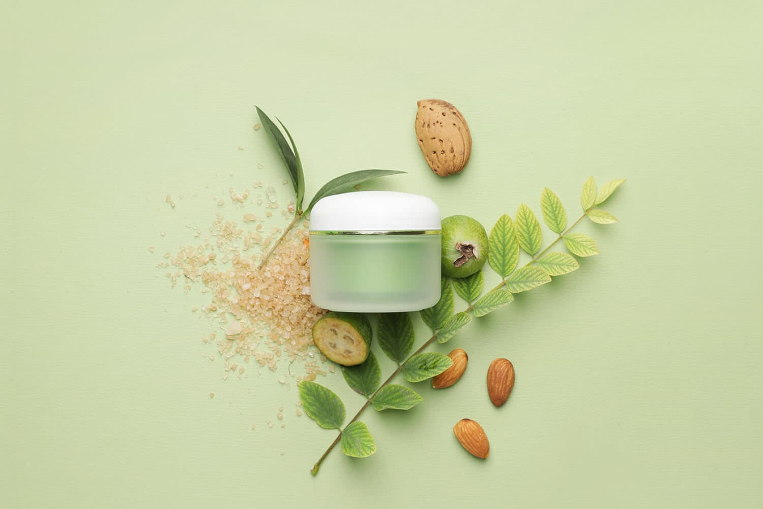 Unlabelled frosted green cream jar on a pale green background, surrounded by almonds, feijoas, leaves and coarse salt. Unlabelled frosted green cream jar on a pale green background, surrounded by almonds, feijoas, leaves and coarse salt.