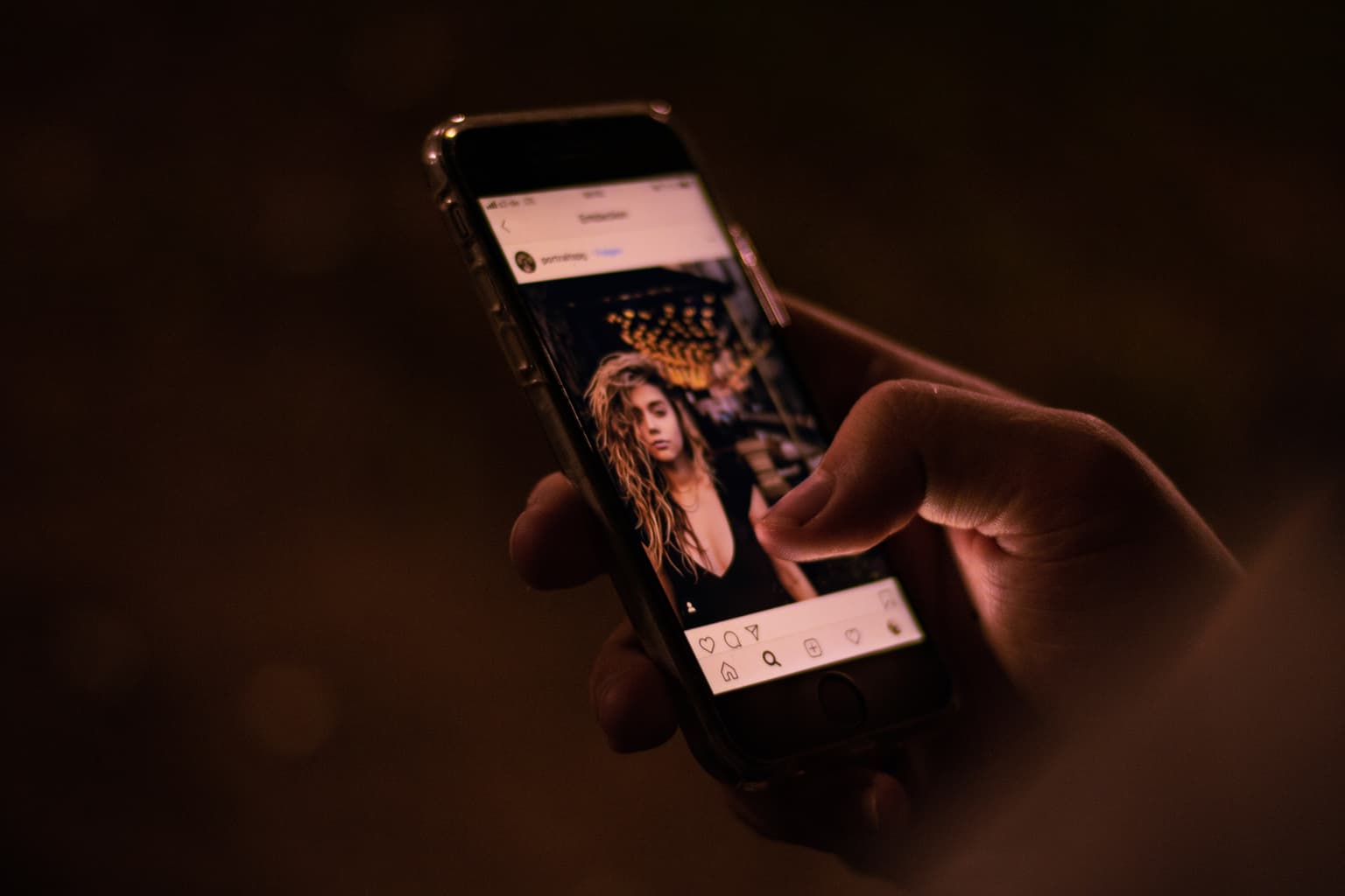 Hand holding an iPhone in dim light, scrolling an Instagram post of a curly-haired woman in a black top. Hand holding an iPhone in dim light, scrolling an Instagram post of a curly-haired woman in a black top.