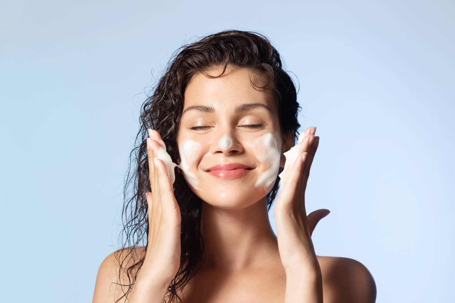 Smiling woman with wet dark curly hair massaging white cleansing foam onto her cheeks against a pale blue background. Smiling woman with wet dark curly hair massaging white cleansing foam onto her cheeks against a pale blue background.