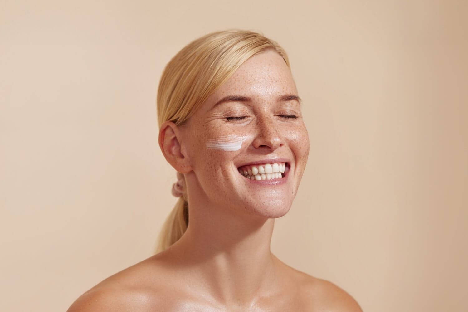 Smiling blonde woman with freckles and closed eyes, a stripe of white cream on her cheek, against a beige backdrop. Smiling blonde woman with freckles and closed eyes, a stripe of white cream on her cheek, against a beige backdrop.