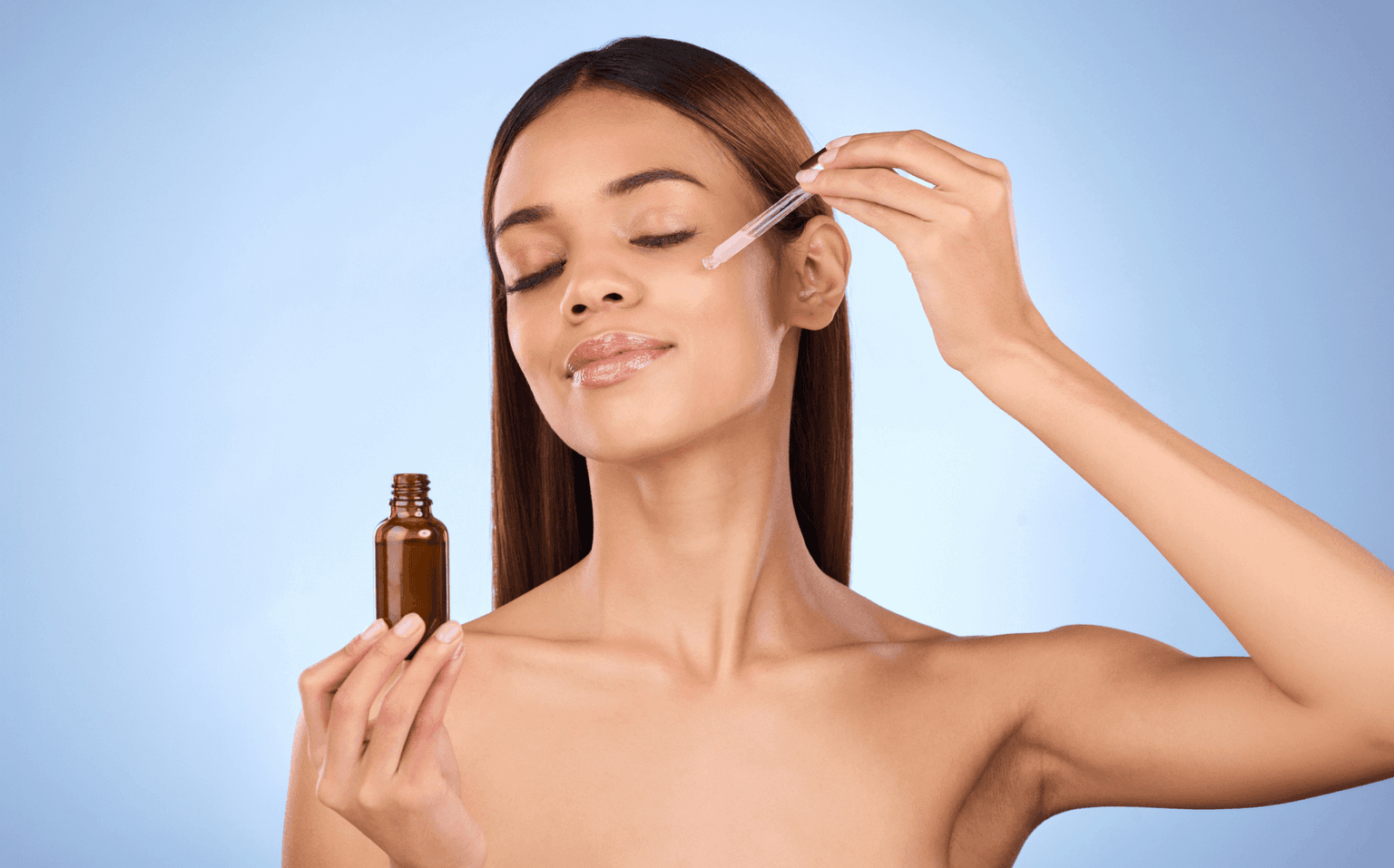 Woman with eyes closed applying a drop from a glass dropper to her cheek, holding an unlabelled amber bottle against a blue backdrop. Woman with eyes closed applying a drop from a glass dropper to her cheek, holding an unlabelled amber bottle against a blue backdrop.