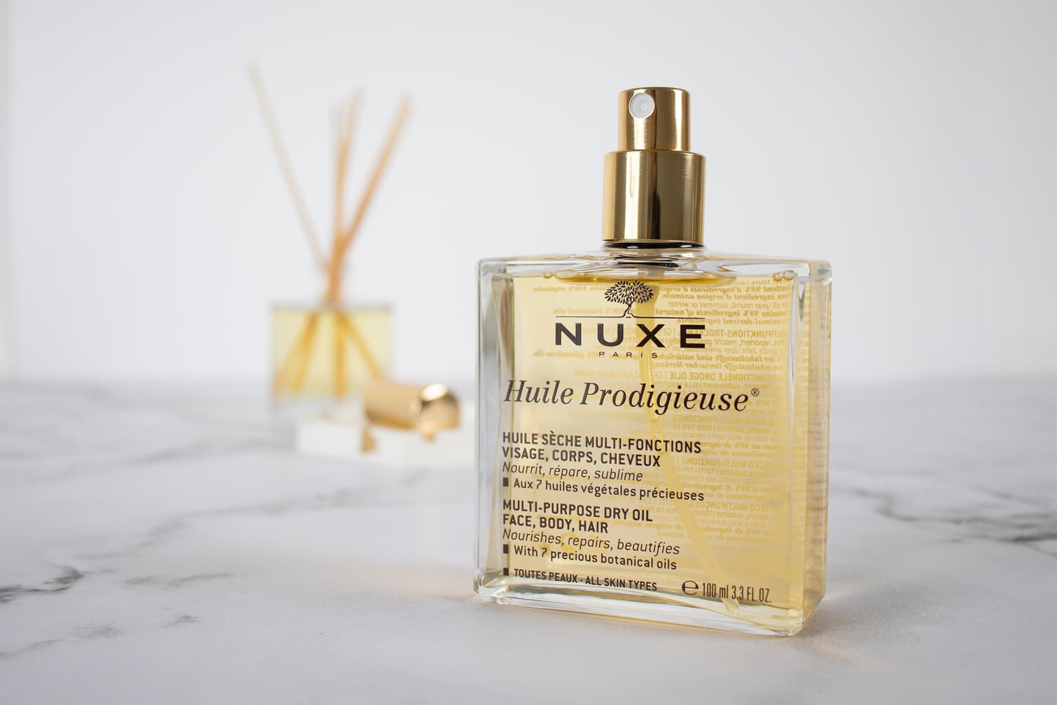 NUXE Paris Huile Prodigieuse multi-purpose dry oil, 100 ml glass bottle with gold spray pump, on marble beside a reed diffuser. NUXE Paris Huile Prodigieuse multi-purpose dry oil, 100 ml glass bottle with gold spray pump, on marble beside a reed diffuser.