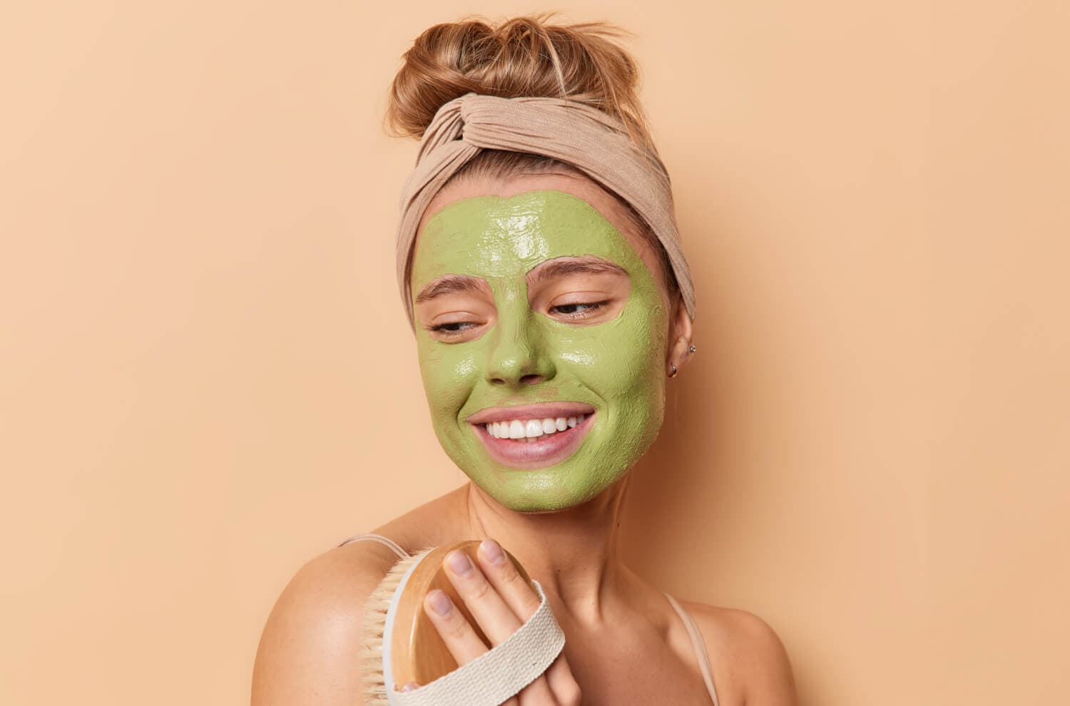 Smiling woman in a beige headband wearing a green clay face mask, holding a wooden natural-bristle body brush against a beige background. Smiling woman in a beige headband wearing a green clay face mask, holding a wooden natural-bristle body brush against a beige background.