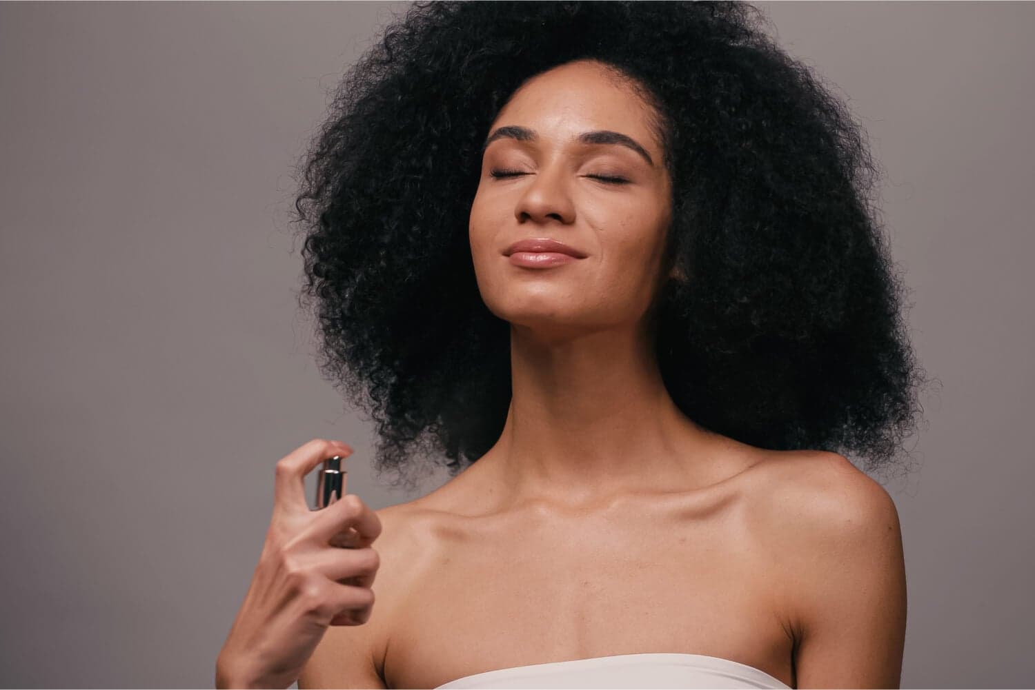 Woman with voluminous curly hair, eyes closed and smiling, spraying an unbranded silver perfume atomiser towards her neck. Woman with voluminous curly hair, eyes closed and smiling, spraying an unbranded silver perfume atomiser towards her neck.