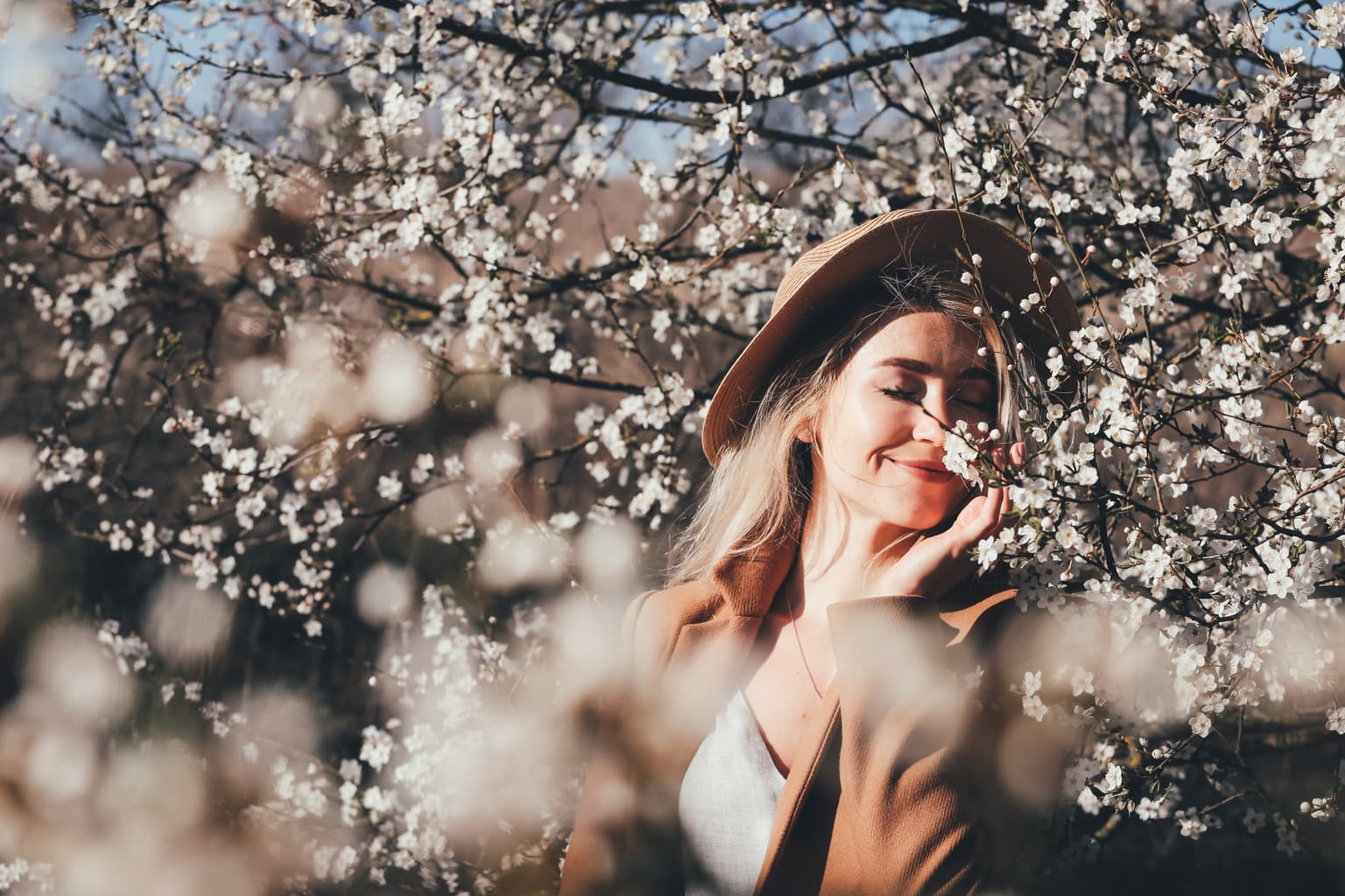 Young woman in a straw hat and tan cardigan smiling with eyes closed as she leans into a branch of white blossom. Young woman in a straw hat and tan cardigan smiling with eyes closed as she leans into a branch of white blossom.