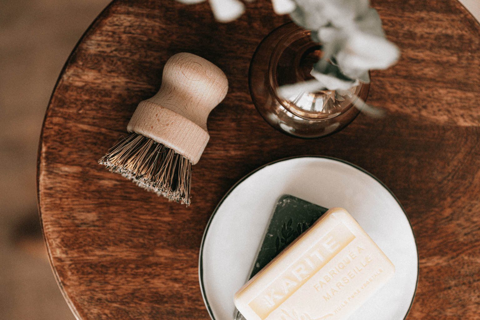 Wooden bristle brush beside a white dish holding a green soap and a cream Karité Fabriqué à Marseille bar on a dark wood table. Wooden bristle brush beside a white dish holding a green soap and a cream Karité Fabriqué à Marseille bar on a dark wood table.
