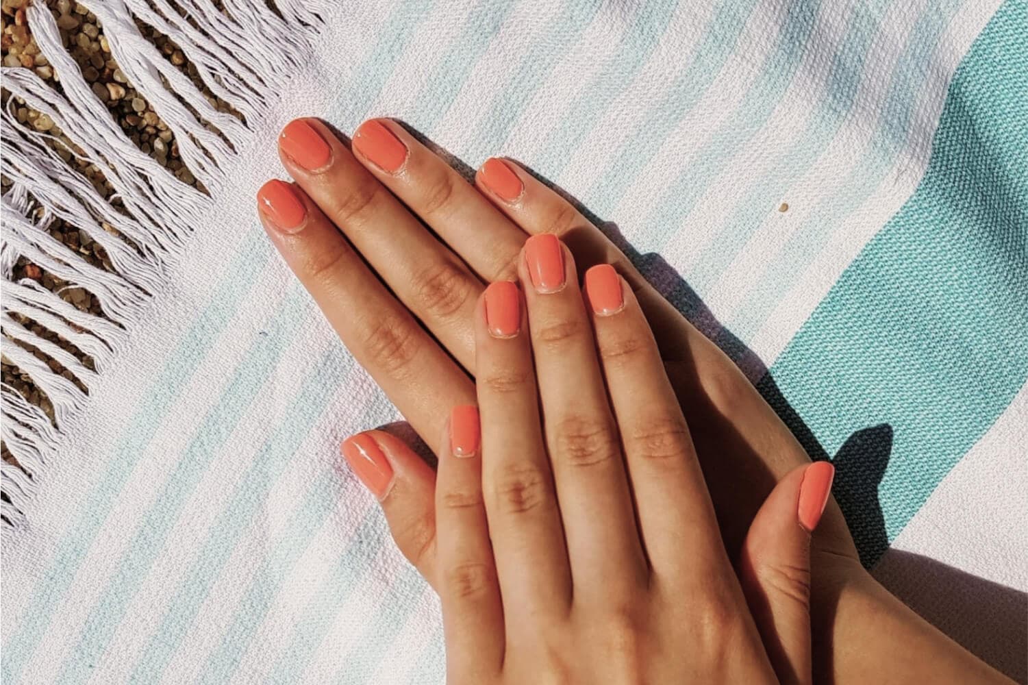 Two hands with short square nails painted glossy coral, resting on a fringed blue and white striped beach towel in the sun.