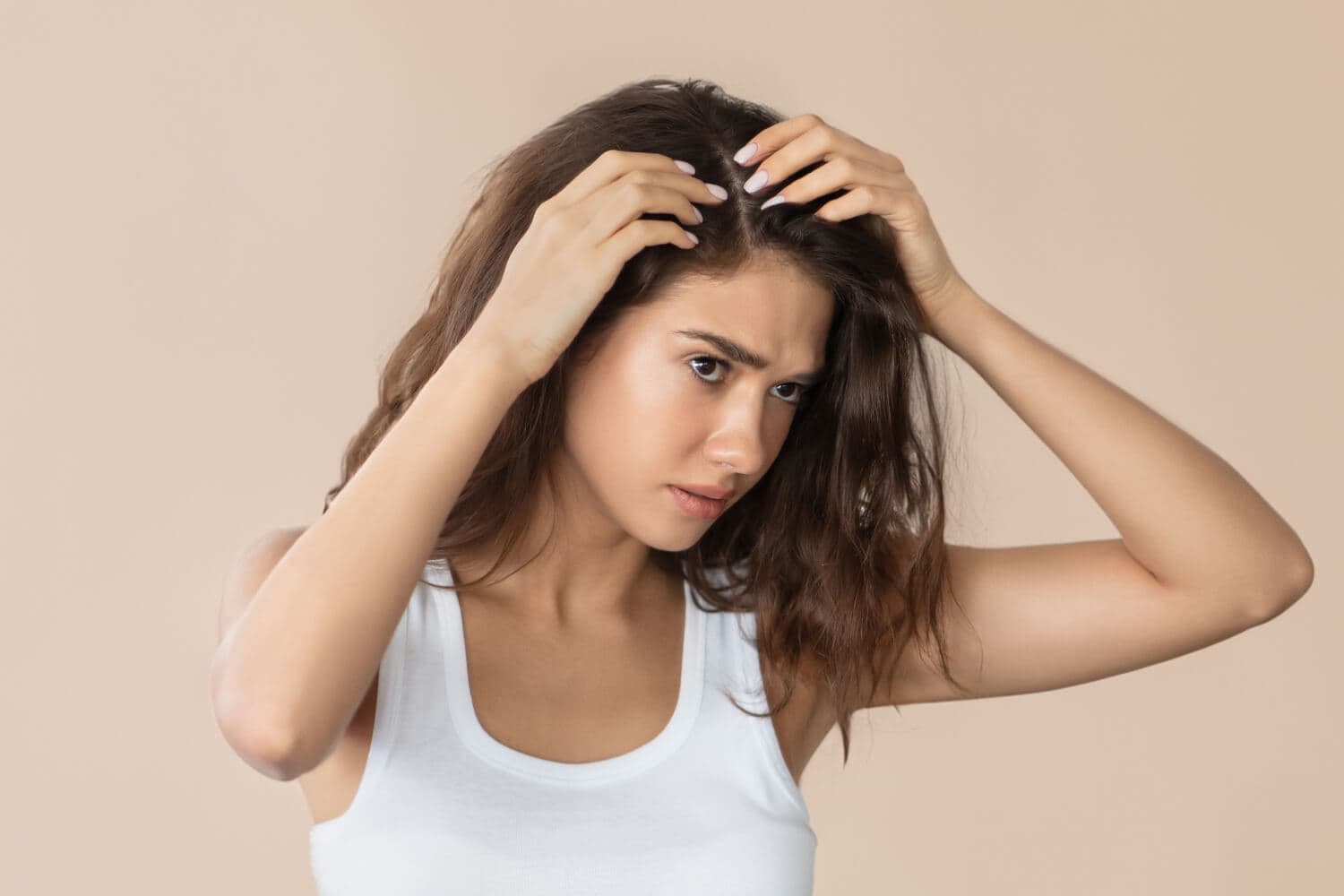 Frowning woman in a white tank top parting her long brown hair with both hands to inspect her scalp, against a beige backdrop. Frowning woman in a white tank top parting her long brown hair with both hands to inspect her scalp, against a beige backdrop.