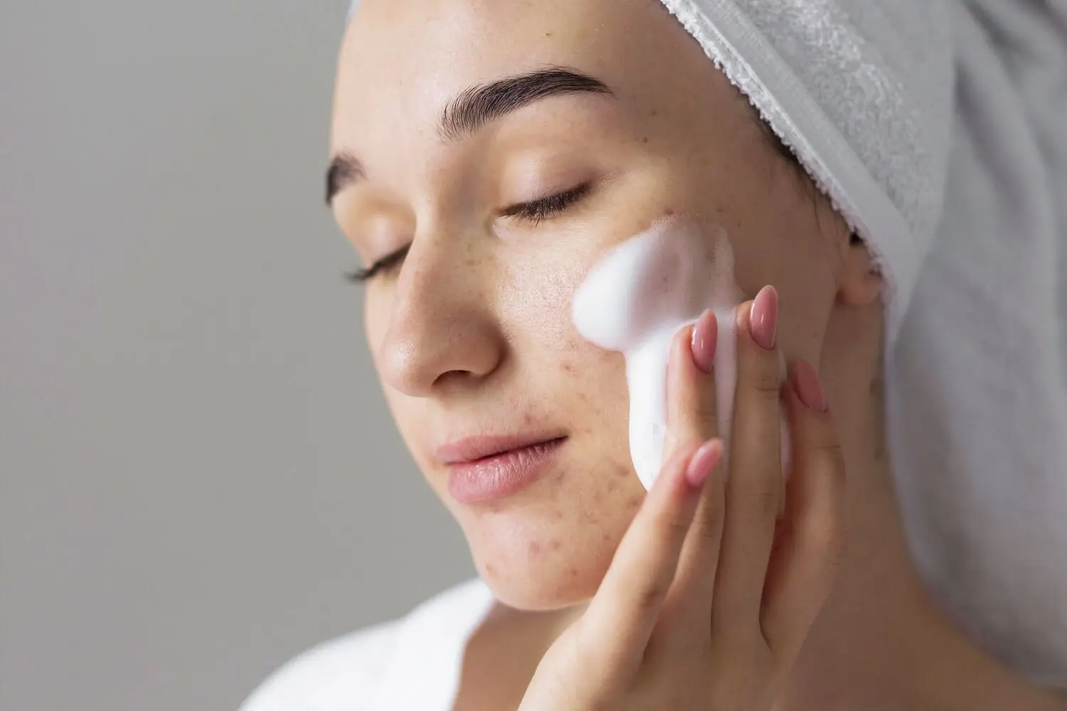 Woman with a white towel turban applying foaming cleanser to her cheek, blemishes and freckles visible on her skin. Woman with a white towel turban applying foaming cleanser to her cheek, blemishes and freckles visible on her skin.