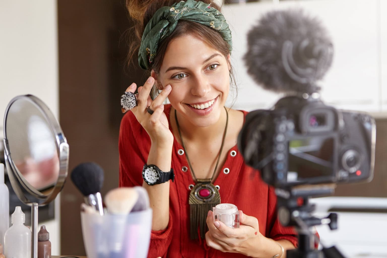 Smiling beauty vlogger in a red top and green headscarf dabs cream under her eye, holding an unlabelled jar to a tripod camera. Smiling beauty vlogger in a red top and green headscarf dabs cream under her eye, holding an unlabelled jar to a tripod camera.