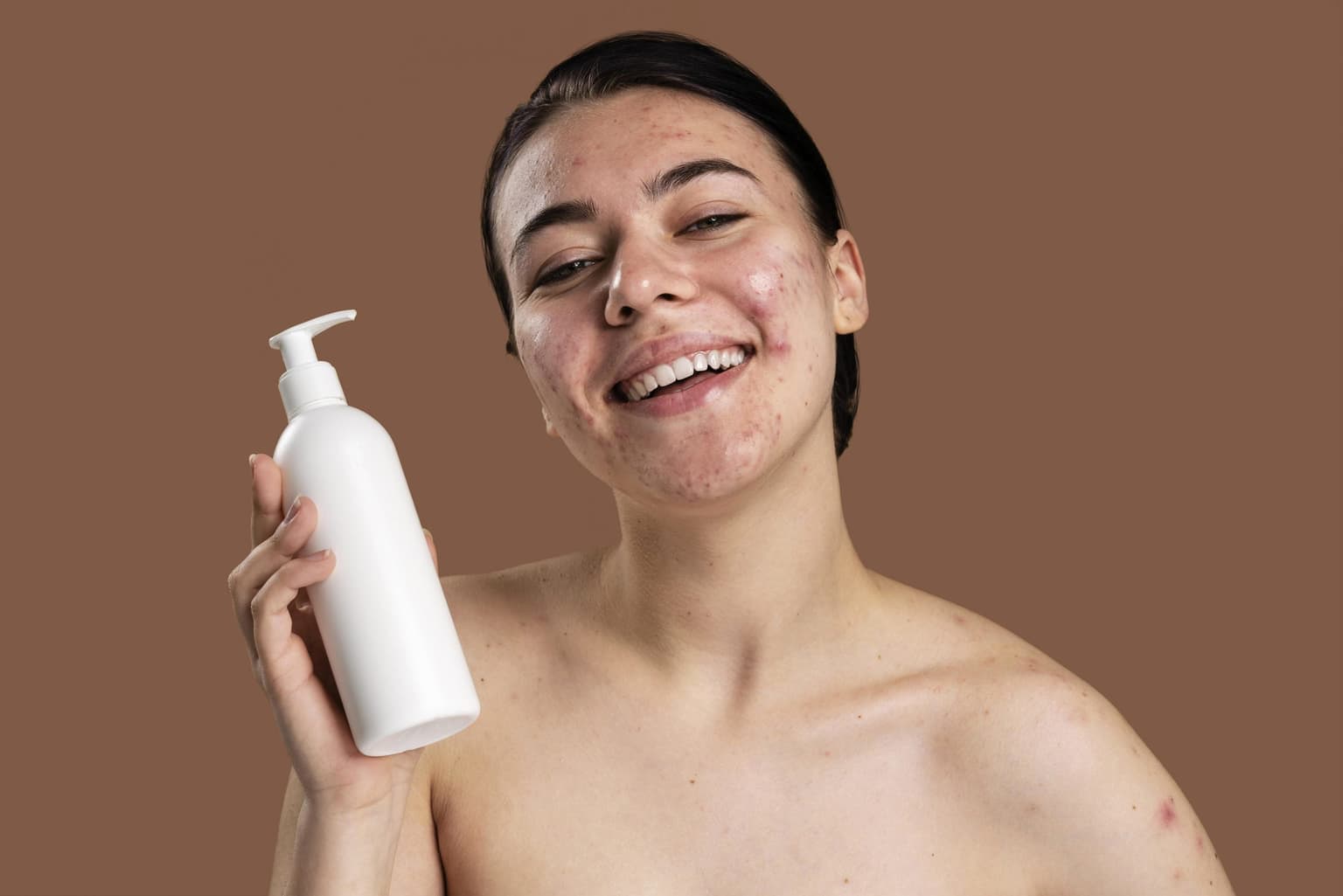 Smiling person with acne-prone skin holding an unlabelled white pump bottle of cleanser against a brown backdrop. Smiling person with acne-prone skin holding an unlabelled white pump bottle of cleanser against a brown backdrop.