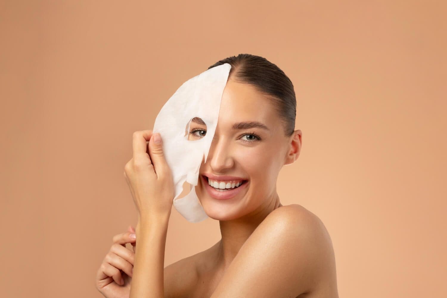 Smiling woman with hair pulled back holding a white sheet mask over half of her face, against a beige backdrop. Smiling woman with hair pulled back holding a white sheet mask over half of her face, against a beige backdrop.