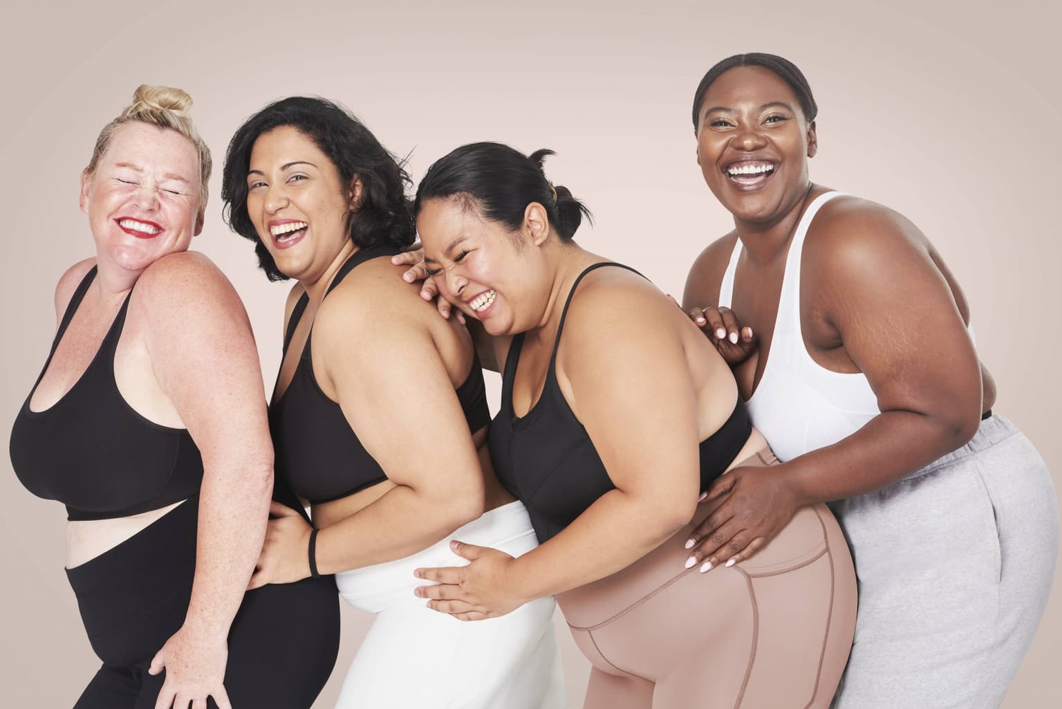 Four laughing women of different ages, sizes and skin tones lean into each other in sports bras and leggings against a soft pink backdrop. Four laughing women of different ages, sizes and skin tones lean into each other in sports bras and leggings against a soft pink backdrop.