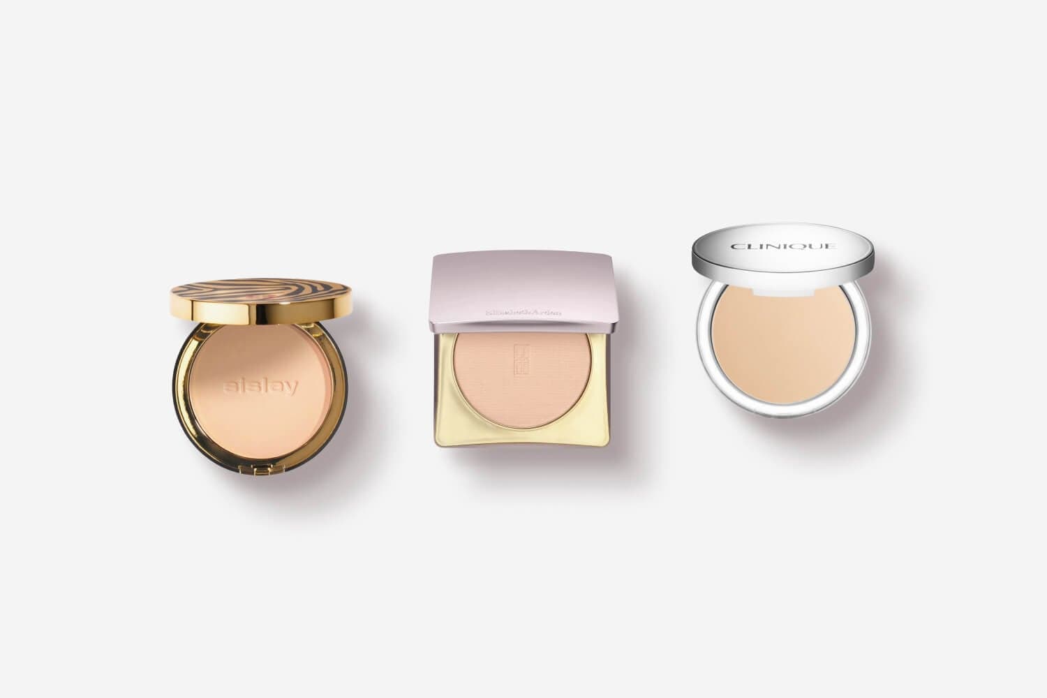 Three open powder compacts on grey: a gold Sisley round compact, a square Elizabeth Arden compact, and a white Clinique compact. Three open powder compacts on grey: a gold Sisley round compact, a square Elizabeth Arden compact, and a white Clinique compact.
