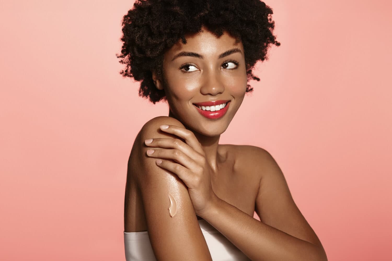 Smiling woman with an afro on a pink backdrop, a swipe of lotion on her glowing bare shoulder and arm. Smiling woman with an afro on a pink backdrop, a swipe of lotion on her glowing bare shoulder and arm.