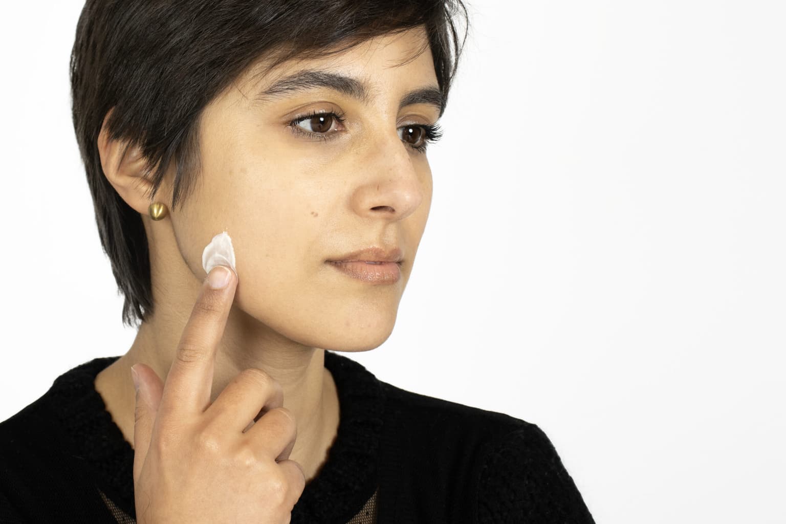 Person with a dark pixie cut dabbing a small dot of white cream onto their jawline with one fingertip, against a white background. Person with a dark pixie cut dabbing a small dot of white cream onto their jawline with one fingertip, against a white background.