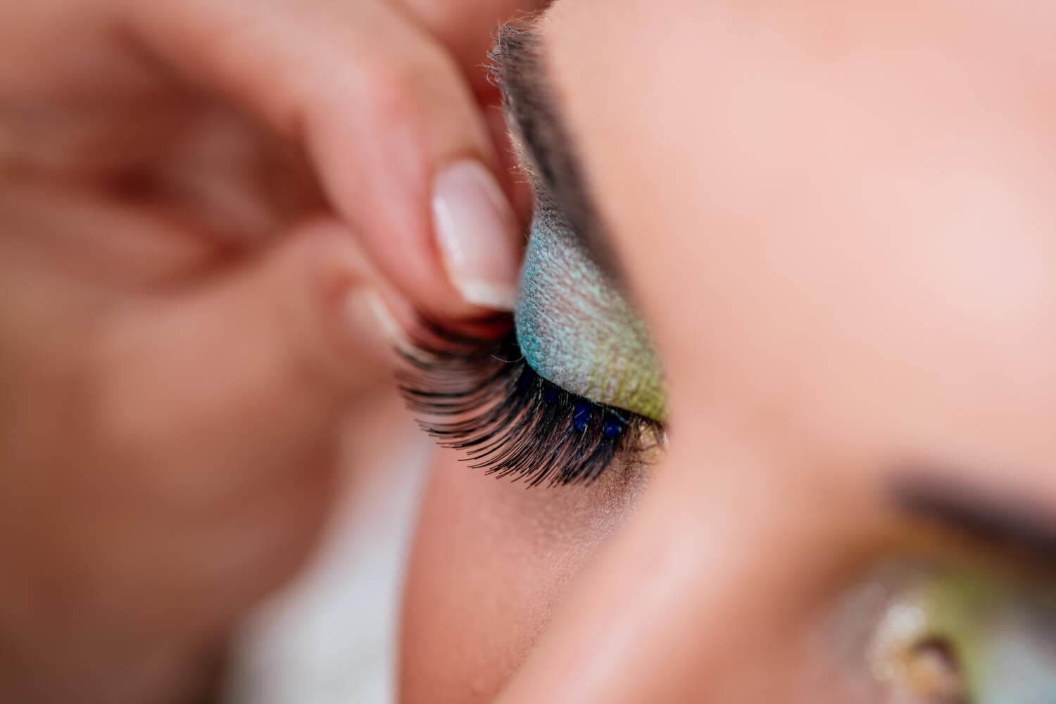 Fingers pressing a strip of false lashes onto a closed eyelid with teal and green shadow and tiny blue gems. Fingers pressing a strip of false lashes onto a closed eyelid with teal and green shadow and tiny blue gems.