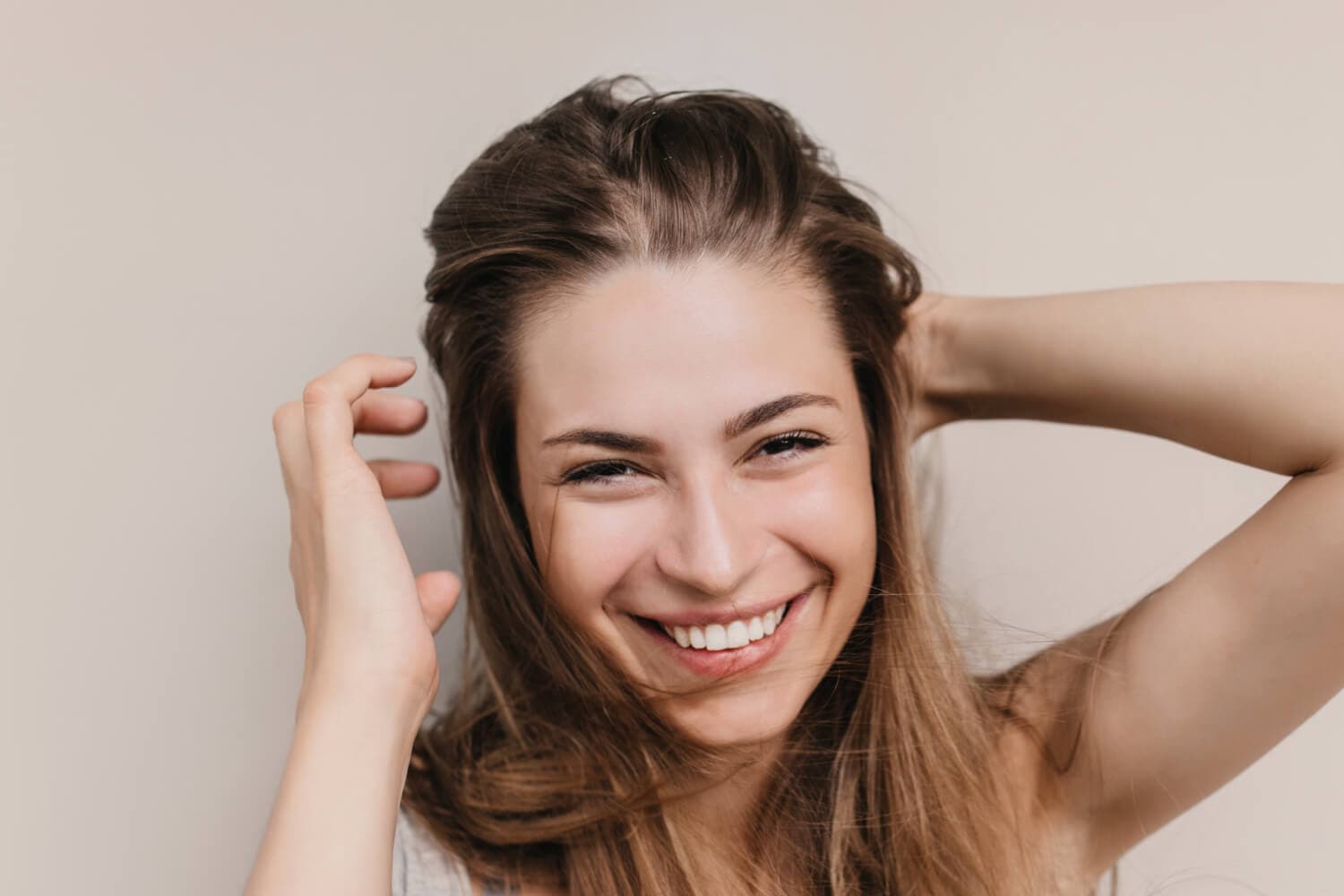 Smiling woman tousling her long light-brown hair with both hands against a plain beige background. Smiling woman tousling her long light-brown hair with both hands against a plain beige background.