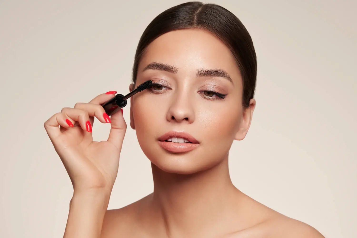 Woman with slicked-back hair and coral nails applying a black mascara wand to her upper lashes against a beige backdrop Woman with slicked-back hair and coral nails applying a black mascara wand to her upper lashes against a beige backdrop