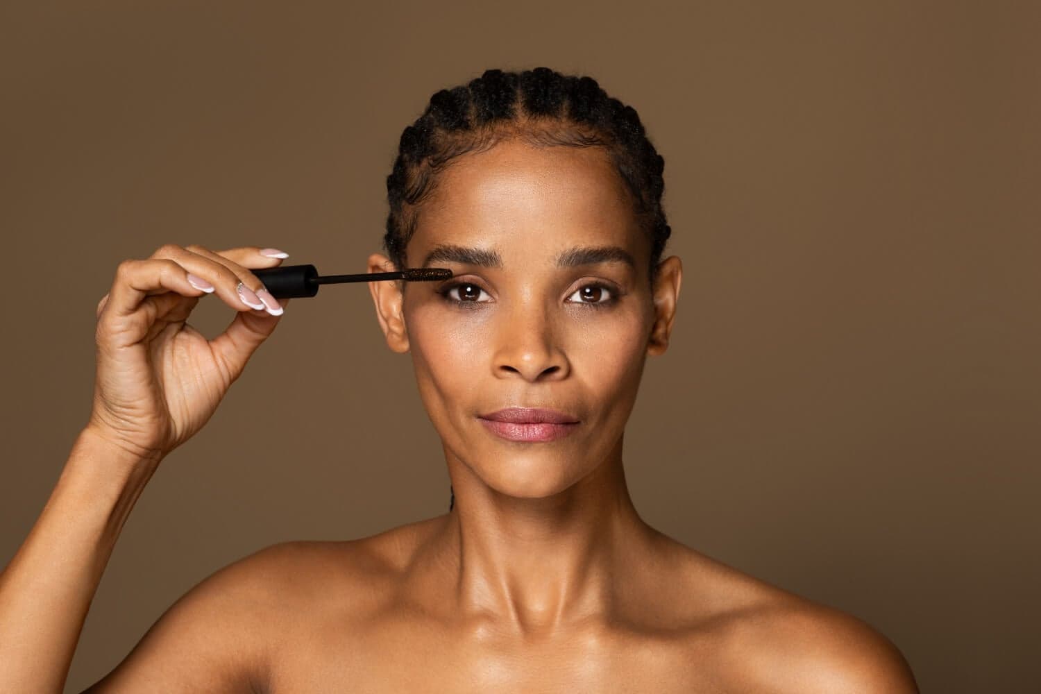 Woman with cornrow braids holding an unbranded black mascara wand with a brown-coated brush to her lashes.