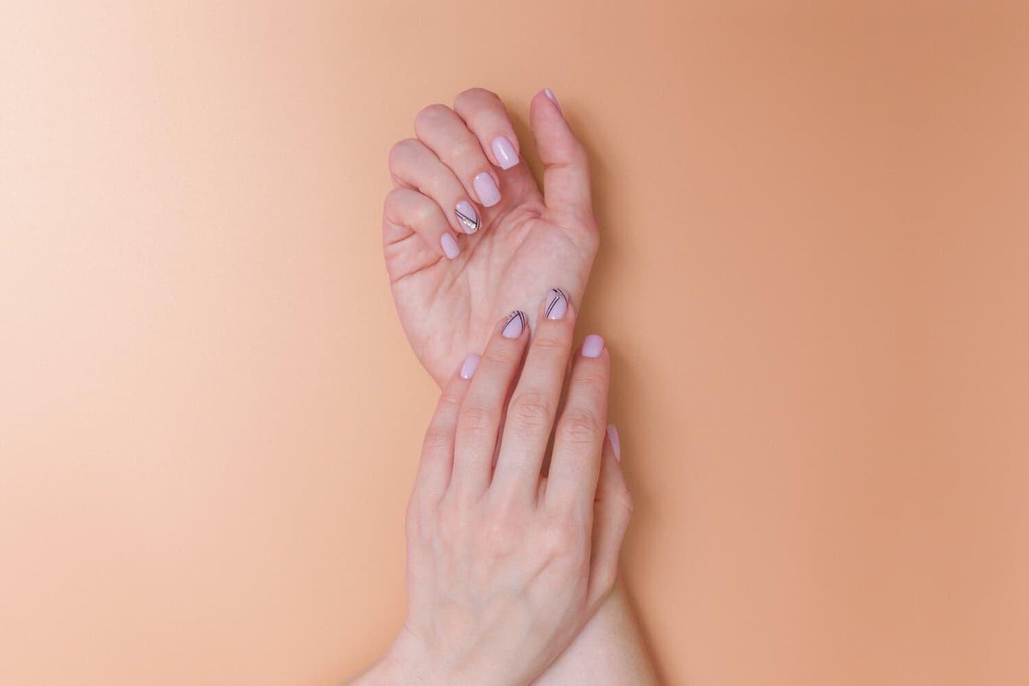 Two hands with short square pale pink nails, two accent nails with fine black lines and tiny gems, on a peach background. Two hands with short square pale pink nails, two accent nails with fine black lines and tiny gems, on a peach background.