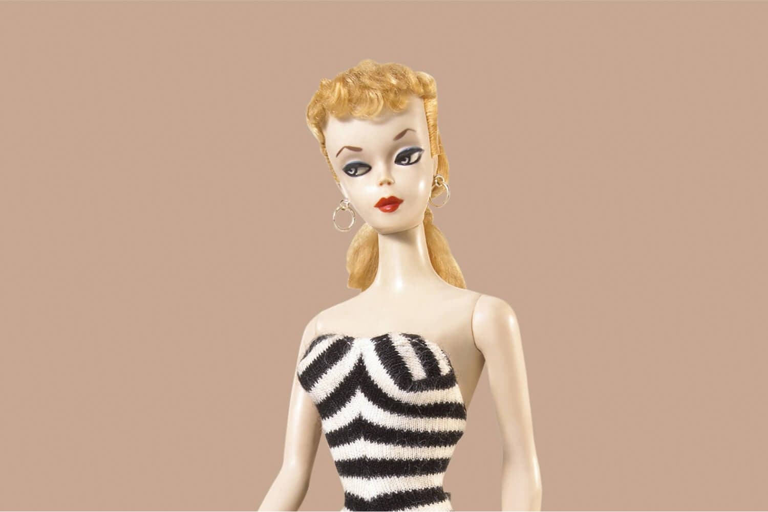 Vintage blonde ponytail doll in a black-and-white striped strapless swimsuit, red lips, winged liner and hoop earrings, on a beige background. Vintage blonde ponytail doll in a black-and-white striped strapless swimsuit, red lips, winged liner and hoop earrings, on a beige background.