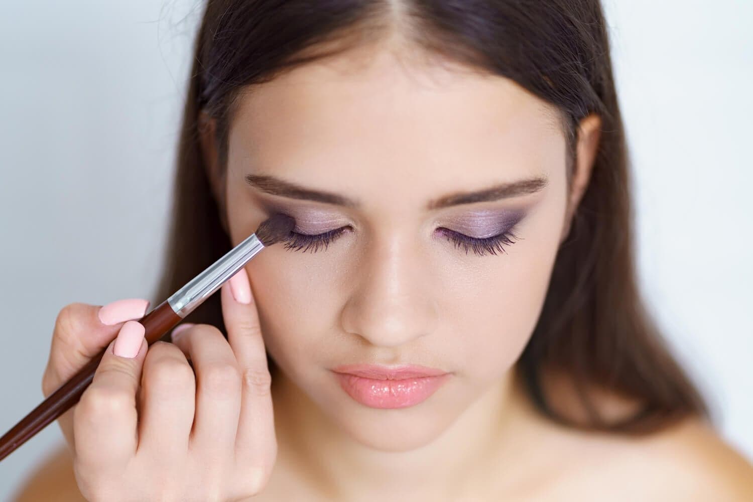 Dark-haired woman with eyes closed as a wooden flat brush blends lilac shimmer along her lash line. Dark-haired woman with eyes closed as a wooden flat brush blends lilac shimmer along her lash line.