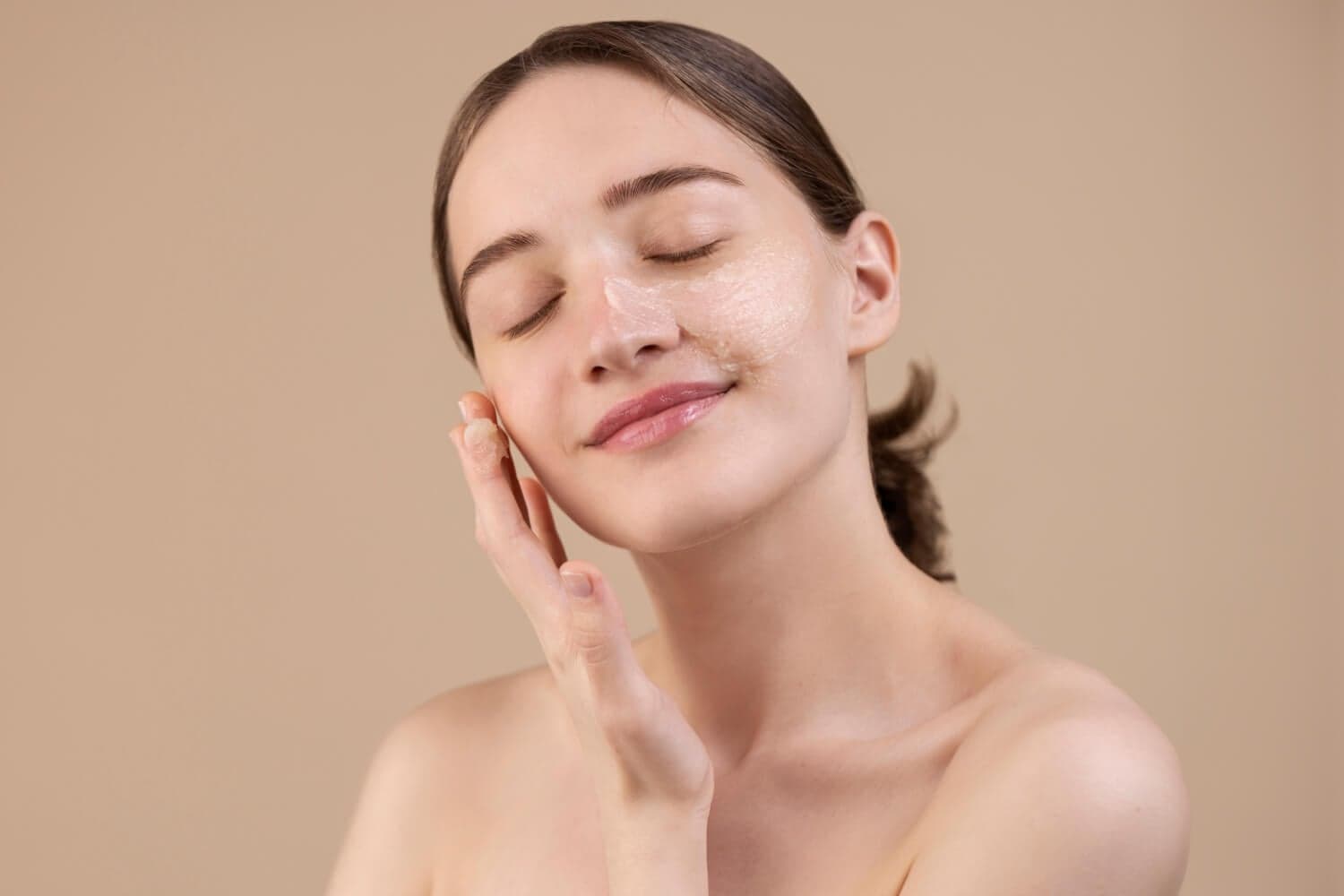 Woman with eyes closed smoothing a streak of white cream across her cheek and nose against a beige backdrop. Woman with eyes closed smoothing a streak of white cream across her cheek and nose against a beige backdrop.