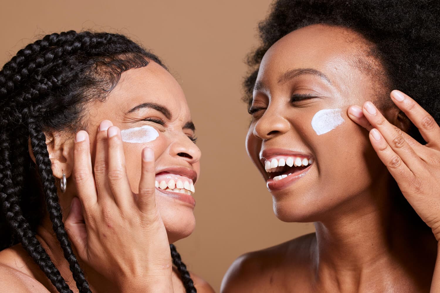 Two laughing women, one with braids and one with an afro, smoothing white cream onto each other's cheeks against a beige backdrop. Two laughing women, one with braids and one with an afro, smoothing white cream onto each other's cheeks against a beige backdrop.