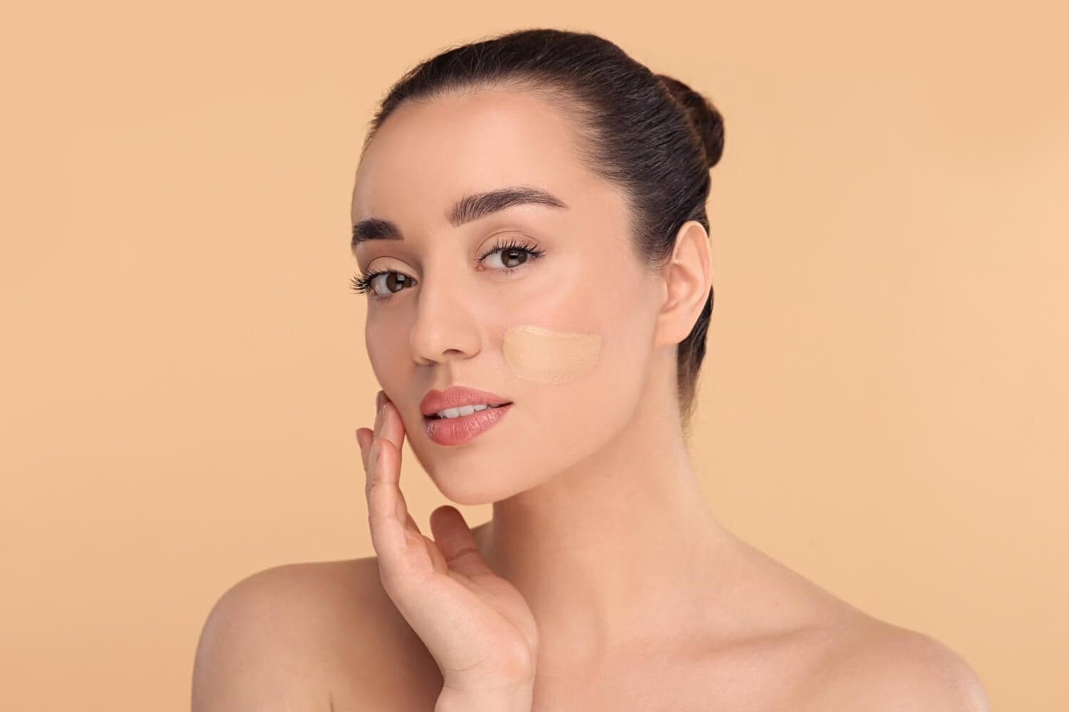 Woman with hair in a bun touching her face, a swatch of liquid foundation blended across her cheek on a beige background. Woman with hair in a bun touching her face, a swatch of liquid foundation blended across her cheek on a beige background.