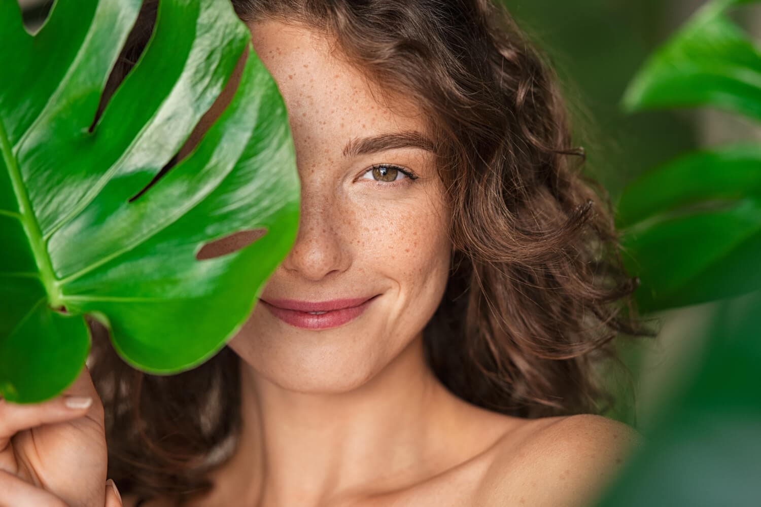 Smiling freckled woman with curly brown hair holding a large green monstera leaf over half of her bare face. Smiling freckled woman with curly brown hair holding a large green monstera leaf over half of her bare face.