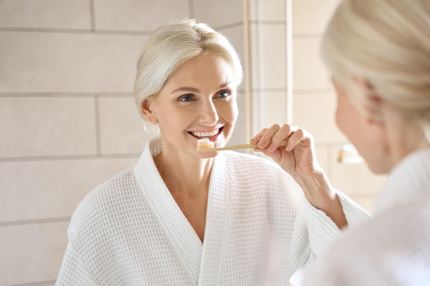 Smiling blonde woman in a white waffle robe holding a bamboo toothbrush with toothpaste, reflected in a bathroom mirror. Smiling blonde woman in a white waffle robe holding a bamboo toothbrush with toothpaste, reflected in a bathroom mirror.