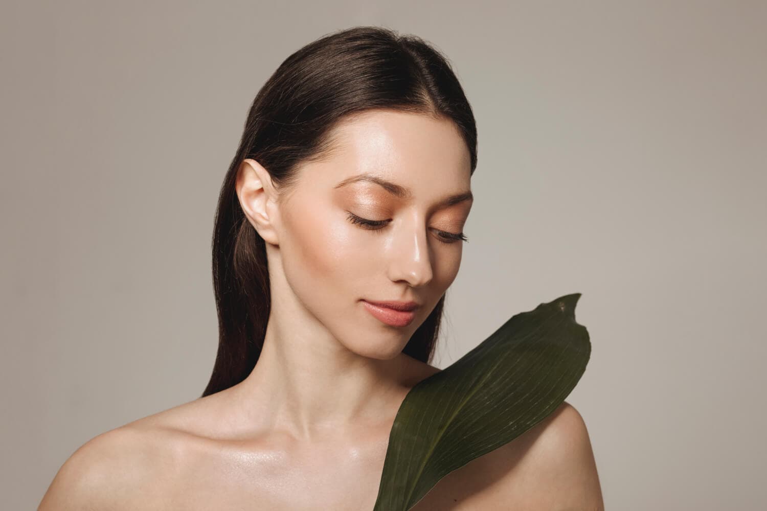 Bare-shouldered model with dewy skin and eyes closed, holding a large dark green leaf against her shoulder on a grey backdrop Bare-shouldered model with dewy skin and eyes closed, holding a large dark green leaf against her shoulder on a grey backdrop