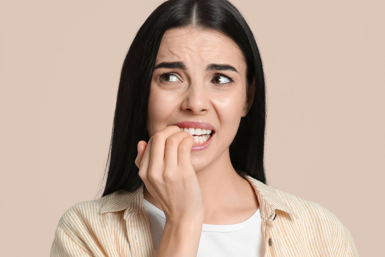 Anxious dark-haired woman in a striped yellow shirt chewing her fingernails against a beige background Anxious dark-haired woman in a striped yellow shirt chewing her fingernails against a beige background