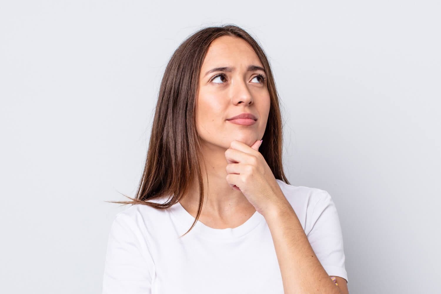 Woman in a white T-shirt with long brown hair, hand on her chin, looking up doubtfully against a plain grey background. Woman in a white T-shirt with long brown hair, hand on her chin, looking up doubtfully against a plain grey background.