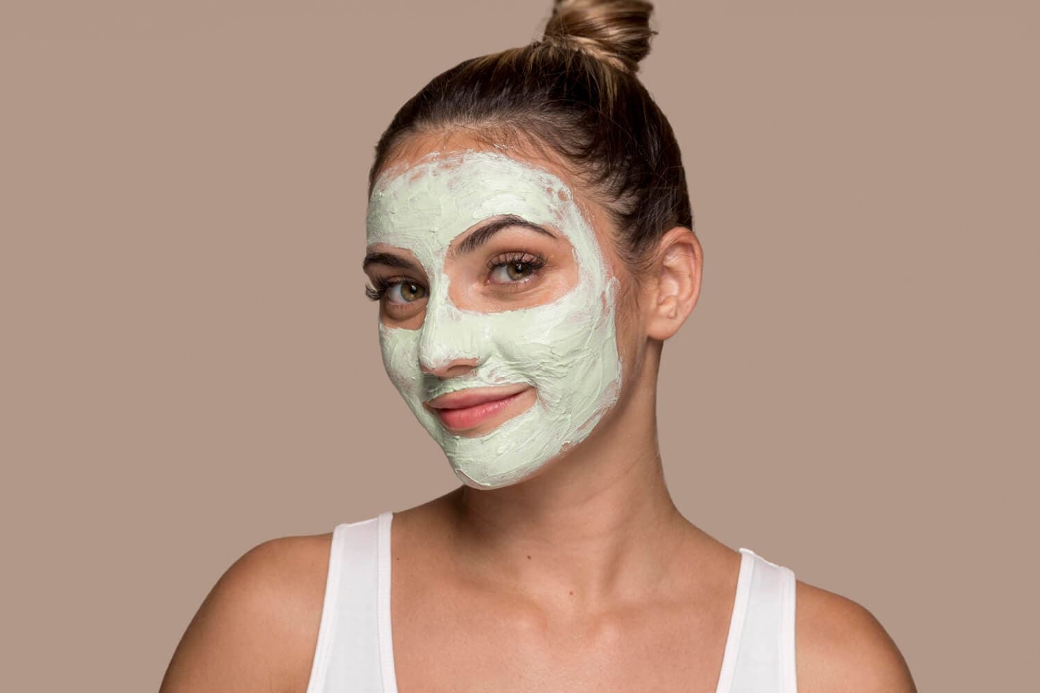 Smiling woman with hair in a bun and a white tank top, wearing a thick pale green clay mask on her face. Smiling woman with hair in a bun and a white tank top, wearing a thick pale green clay mask on her face.