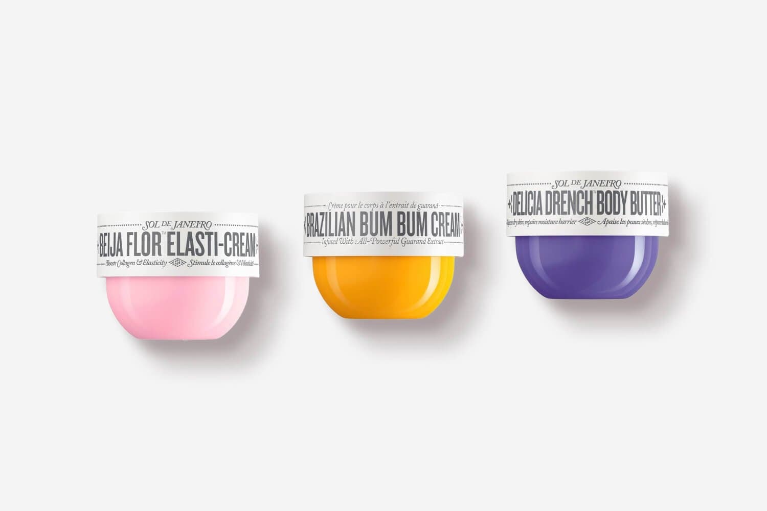 Three Sol de Janeiro jars in a row: pink Beija Flor Elasti-Cream, yellow Brazilian Bum Bum Cream and purple Delicia Drench Body Butter. Three Sol de Janeiro jars in a row: pink Beija Flor Elasti-Cream, yellow Brazilian Bum Bum Cream and purple Delicia Drench Body Butter.