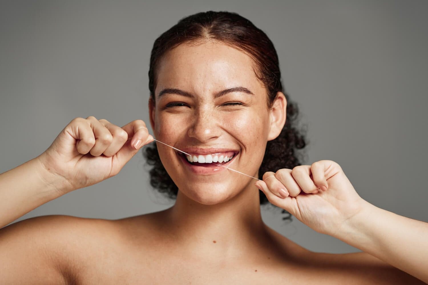 Smiling woman with freckles and dark curly hair holding a strand of dental floss taut against her teeth, on a grey background. Smiling woman with freckles and dark curly hair holding a strand of dental floss taut against her teeth, on a grey background.