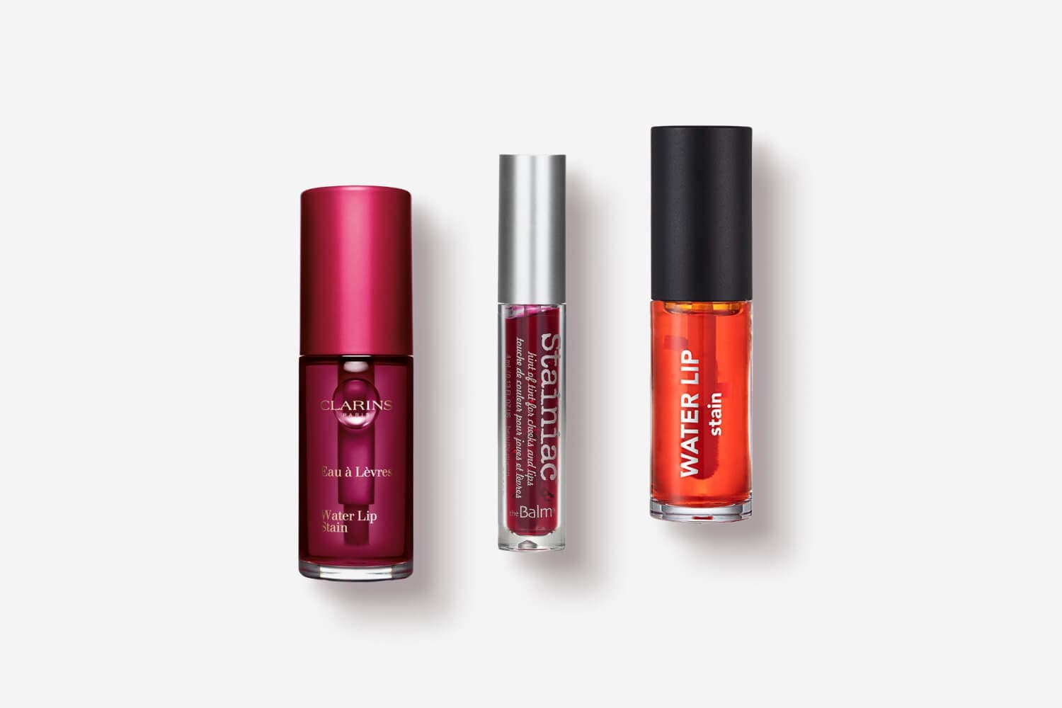 Clarins Eau à Lèvres Water Lip Stain, theBalm Stainiac lip and cheek tint, and an orange Water Lip Stain lined up on white. Clarins Eau à Lèvres Water Lip Stain, theBalm Stainiac lip and cheek tint, and an orange Water Lip Stain lined up on white.